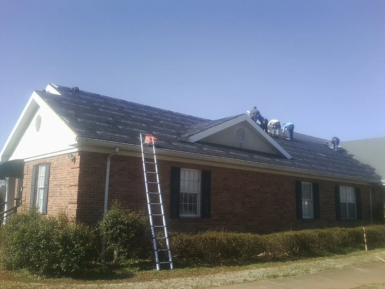 Crew tearing shingles off a brick ranch roof with a ladder set — Emergency Roof Repair work by our crew in Munnsville