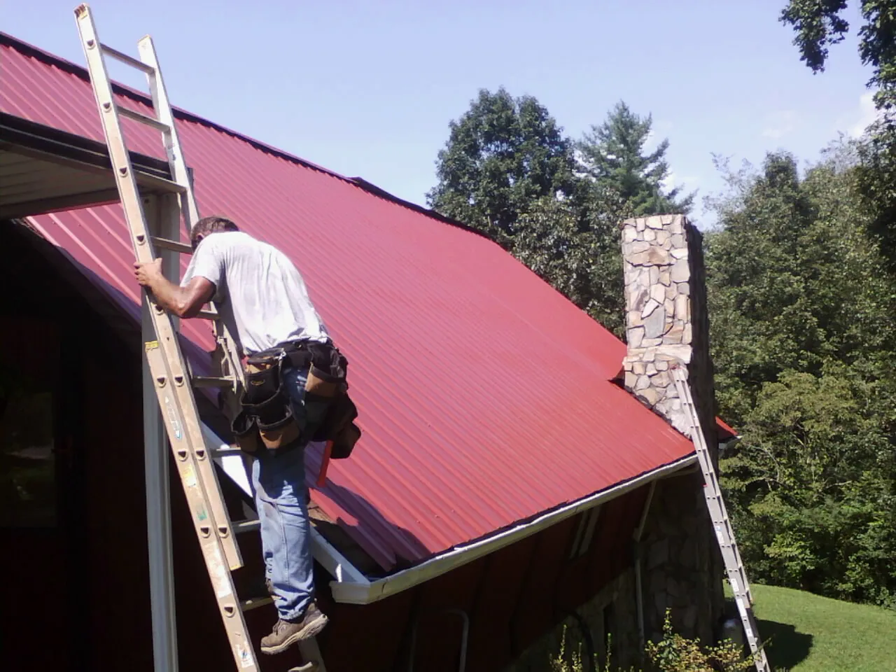 Roofer on a ladder beside a red metal roof and stone chimney — Soffit & Fascia Repair by Roofing Brothers in Munnsville